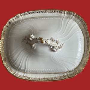 May include: An oval, white porcelain serving dish with a lid. The dish has a gold-coloured, ornate trim and a decorative handle with floral and berry accents. The lid features a wavy, textured design.