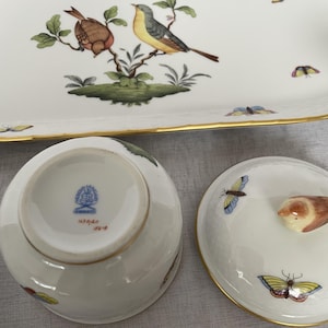 May include: A white porcelain serving set with gold trim, featuring hand-painted birds and butterflies. The set includes a rectangular tray and a covered bowl. The bowl's lid has a bird-shaped handle. The underside of the bowl has a blue stamp.