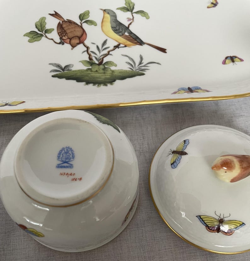 May include: A white porcelain serving set with gold trim, featuring hand-painted birds and butterflies. The set includes a rectangular tray and a covered bowl. The bowl's lid has a bird-shaped handle. The underside of the bowl has a blue stamp.