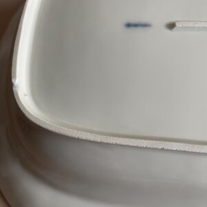 May include: A white, rectangular ceramic serving platter with a slightly curved edge. The underside of the platter has a textured, unglazed rim. A small blue marking is visible on the surface.