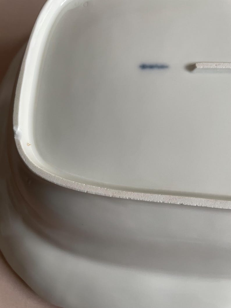 May include: A white, rectangular ceramic serving platter with a slightly curved edge. The underside of the platter has a textured, unglazed rim. A small blue marking is visible on the surface.