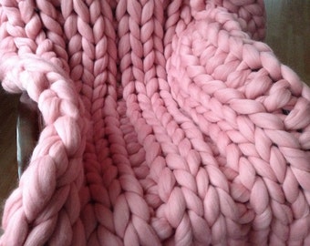 May include: A soft, pink chunky knit blanket made with thick yarn. The blanket is folded and draped over a chair.