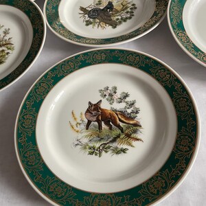 May include: A collection of white ceramic plates with a green and gold decorative border. Each plate features a different wildlife scene in the center, including a fox, birds, and foliage. The plates are arranged on a white surface.