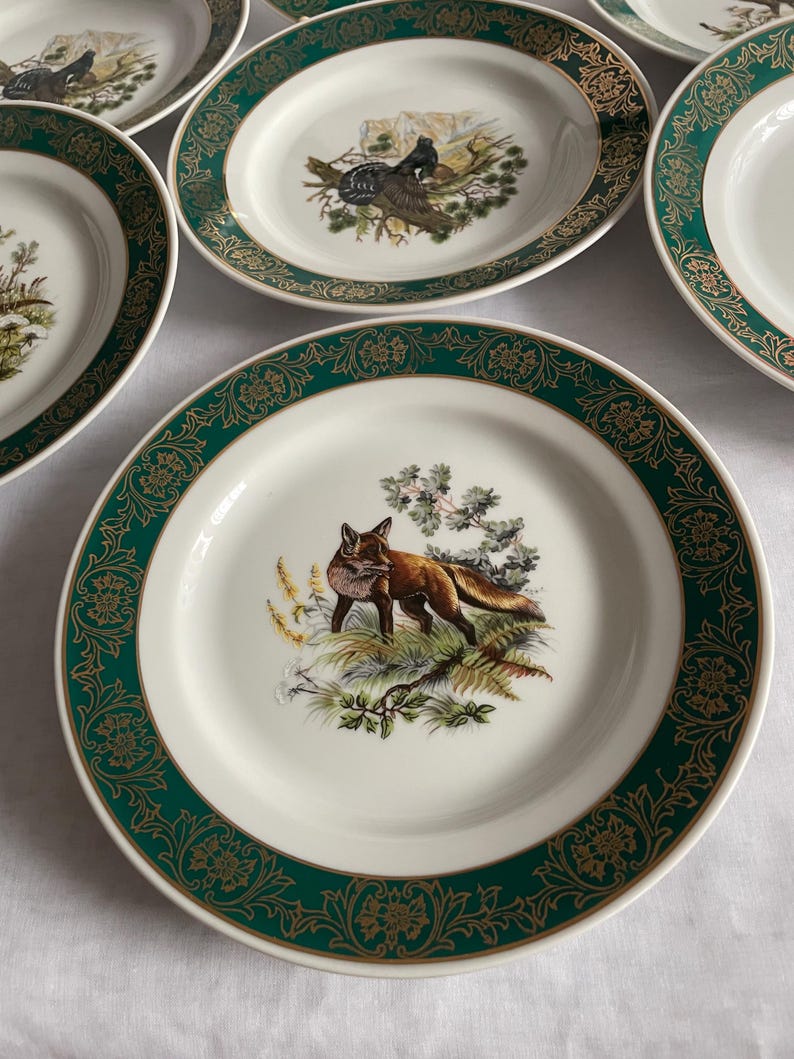 May include: A collection of white ceramic plates with a green and gold decorative border. Each plate features a different wildlife scene in the center, including a fox, birds, and foliage. The plates are arranged on a white surface.