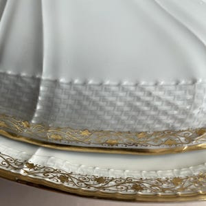 May include: Close-up of a white porcelain serving dish with a lid. The dish features a woven texture around the rim and is accented with gold floral designs. The lid has a fluted design.