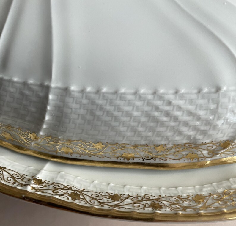 May include: Close-up of a white porcelain serving dish with a lid. The dish features a woven texture around the rim and is accented with gold floral designs. The lid has a fluted design.
