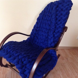 May include: A blue knitted throw blanket draped over a wooden rocking chair. The blanket is made with thick yarn and has a chunky knit texture.