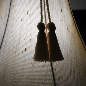 May include: A white lampshade with a textured fabric surface and two white tassels hanging from the top.