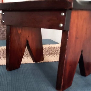 May include: A dark brown wooden stool with a simple design. The stool has a rectangular top and four legs that are shaped like a V. The stool is made of solid wood and has a natural finish.