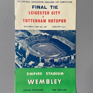May include: Vintage football program for the 1961 Football Association Challenge Cup Final Tie between Leicester City and Tottenham Hotspur. The program features a stadium aerial view, with text in red and black on a cream background, and green at the bottom.