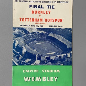 May include: Vintage football program for the 1962 Football Association Challenge Cup Final Tie between Burnley and Tottenham Hotspur. The program features a stadium aerial view, with text in blue, white, and green. The game was held at Wembley Stadium.