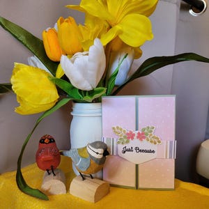 Op de afbeelding: Een decoratieve opstelling met een boeket gele en witte bloemen in een witte pot, twee houten vogels en een wenskaart. De kaart is roze met witte stippen en de tekst "Just Because".