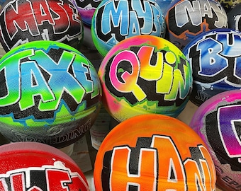 May include: A selection of basketballs, each adorned with graffiti-style lettering in a range of colours. The basketballs display names or words in bold, outlined fonts, with bright shades including blue, green, orange, and pink. Some balls have the Spalding logo.