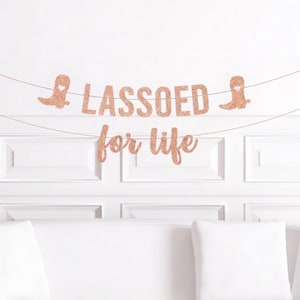 以下が含まれることがあります： 「LASSOED for life」の文字とカウボーイブーツのイラストが描かれた、ローズゴールドのグリッターバナー。白い壁を背景に、白いクッションが置かれた白いソファの上に飾られています。