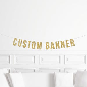 May include: A gold glitter "CUSTOM BANNER" hangs against a white wall. The banner is strung on a thin string. Below the banner is a white sofa with white cushions.