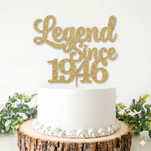 以下が含まれることがあります： 白いフロストケーキの上に「Legend Since 1946」の文字が入った金色のグリッターケーキトッパー。ケーキは背景に緑のある木の台の上にあります。トッパーは誕生日ケーキの装飾要素です。
