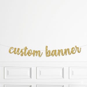 May include: A gold glitter 'custom banner' strung across a white wall. The banner is made of individual cursive letters. The wall has white paneling below the banner.