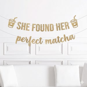 May include: A gold glitter banner with the text "SHE FOUND HER perfect matcha" and two coffee cup illustrations. The banner is strung across a white wall above a white couch with pillows. The banner is a decorative item.