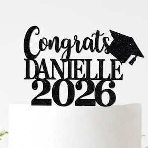 May include: A white cake with a black glitter cake topper that reads "Congrats DANIELLE 2026" and a graduation cap. The cake sits on a wooden base, decorated with greenery.