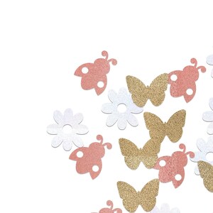 May include: Decorative confetti in the shape of butterflies, ladybirds, and flowers. The confetti is in gold, white, and pink, with a glittery texture. The background is plain white.