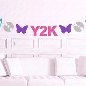 May include: A decorative banner featuring the text Y2K in pink glitter, flanked by purple glitter butterflies, silver glitter compact discs, and teal glitter flip phones. The elements are strung together on a thin white cord for display.