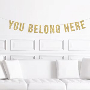 May include: A decorative banner featuring individual gold glitter letters strung together on a thin cord. The letters spell out the phrase YOU BELONG HERE in a bold, sans-serif font. The glitter finish provides a reflective, textured surface.