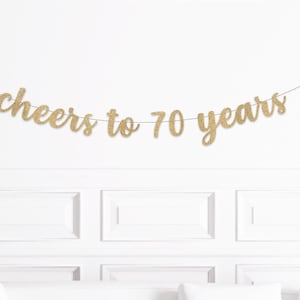May include: A gold glitter banner that reads "cheers to 70 years" is strung across a white wall. The banner is made of gold glitter cardstock and is suspended by a thin, black string. The wall has a decorative panel design.