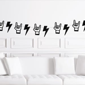 Peut inclure: Une guirlande noire pailletée avec des gestes de la main et des éclairs. La guirlande est tendue sur un mur blanc au-dessus d'un canapé blanc. Les gestes de la main sont le signe du rock and roll.