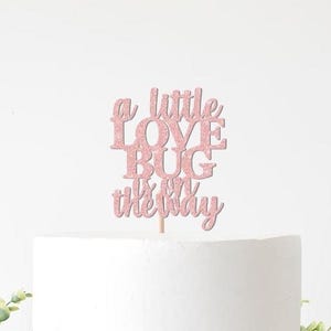 May include: A pink glitter cake topper with the phrase "a little love bug is on the way". The topper is on a white cake, with greenery in the foreground. Perfect for a baby shower.