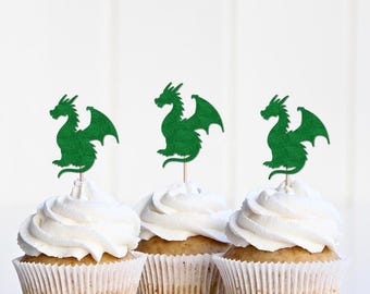 May include: Three cupcakes topped with white frosting and green dragon-shaped toppers. The cupcakes are in white paper liners, and the background is a plain white surface. The dragon toppers are made of a textured green material.