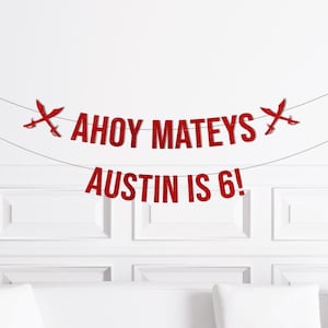 May include: Red glitter banner with the words "AHOY MATEYS" and "AUSTIN IS 6!" strung across a white wall. The banner includes crossed swords on each end. Perfect for a pirate-themed birthday party.