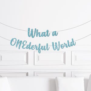 May include: A light blue glitter banner with the words "What a Wonderful World" strung across a white wall. The banner is suspended by thin silver strings, and the text is in a cursive font.