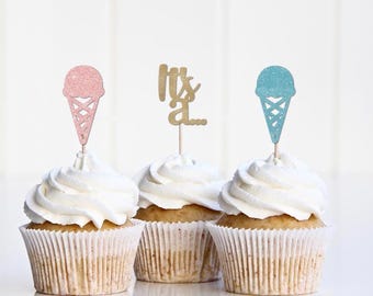 May include: Three cupcakes with white frosting and decorative toppers. One cupcake has a pink glitter ice cream cone topper, another has a gold "It's a..." topper, and the third has a blue glitter ice cream cone topper. The cupcakes are in white paper liners.