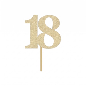 May include: A gold glitter cake topper in the shape of the number "18" on a wooden stick. The numbers are in a bold font and covered in gold glitter. The background is white.