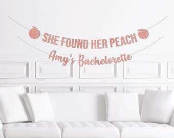 Può includere: Uno striscione rosa glitterato con la scritta "SHE FOUND HER PEACH" e "Amy's Bachelorette" su un muro bianco. Due illustrazioni di pesche affiancano il testo. Lo striscione è sopra un divano bianco.
