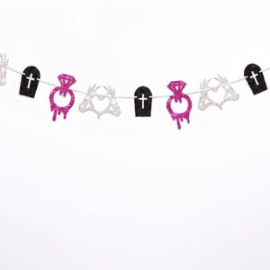May include: A decorative banner featuring alternating glittery cutouts strung on a white cord. The shapes include silver skeleton hands forming heart shapes, black coffins with white cross cutouts, and hot pink diamond rings with a dripping effect.