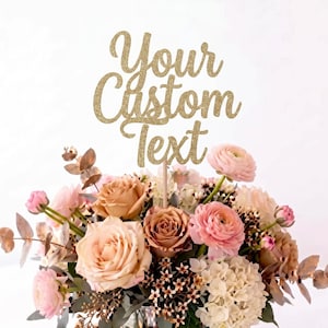 May include: A gold glitter cake topper featuring the text Your Custom Text in a cursive script font, mounted on a thin wooden stick and displayed emerging from a floral arrangement of pink, peach, and white roses and green eucalyptus leaves.