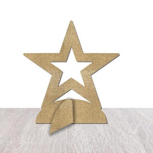 May include: A gold-colored star-shaped decorative object with a star cutout in the center. The star sits on a base with a curved design. The object is made of a textured material and is displayed on a light-colored wooden surface.