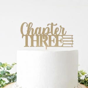 以下が含まれることがあります： 「Chapter THREE」の文字と本のスタックが描かれた、金色のグリッターケーキトッパー。白いケーキの上にトッパーが置かれ、背景には緑の葉があります。