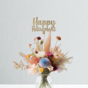 May include: A gold glitter cake topper with the text "Happy Retirement" on a stick, placed in a bouquet of dried flowers in a clear glass vase.