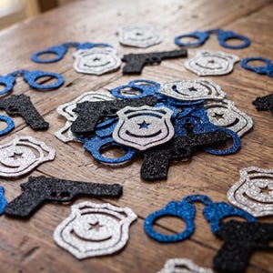 May include: A collection of glittery paper cutouts in the shapes of police badges, handcuffs, and handguns. The badges are silver, the handcuffs are blue, and the handguns are black. These decorative pieces are scattered across the surface.