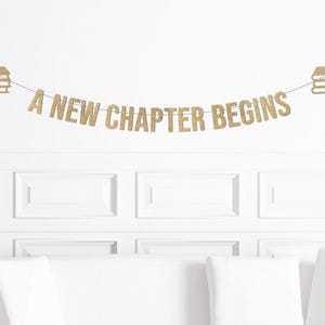 May include: A gold glitter banner with the phrase "A NEW CHAPTER BEGINS" strung across a white wall. The banner is decorated with gold glitter book icons on each end. The banner is hanging above a white couch with white pillows.