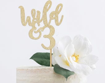 以下が含まれることがあります： 白い花で飾られたケーキに、「Wild & 3」と書かれた金のグリッターケーキトッパー。ケーキトッパーは木の棒に取り付けられています。ケーキは白く、表面はざらざらしています。