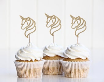 May include: Three cupcakes with white frosting and gold glitter unicorn toppers. The cupcakes are in white paper liners. The background is a plain white surface.