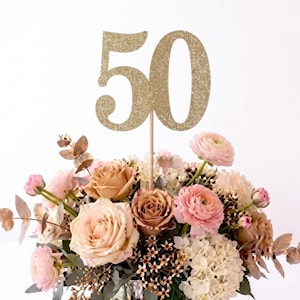 May include: A gold glitter number 50 cake topper attached to a thin wooden stick, positioned above a floral arrangement featuring light pink, peach, and cream-colored roses, ranunculus, and white hydrangeas with green eucalyptus leaves.
