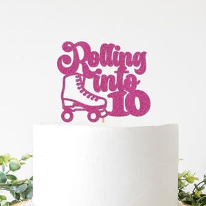 May include: A pink glitter cake topper with the words "Rolling into 10" and a roller skate illustration. The topper is placed on top of a white cake, with greenery in the foreground.