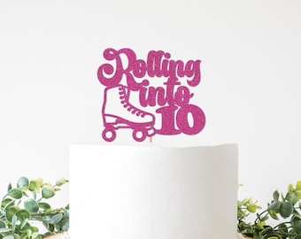 May include: A pink glitter cake topper with the words "Rolling into 10" and a roller skate illustration. The topper is placed on top of a white cake, with greenery in the foreground.