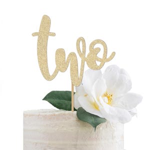May include: A gold glitter "two" cake topper on a white frosted cake, decorated with a white flower and green leaves. The cake topper is attached to a wooden stick.