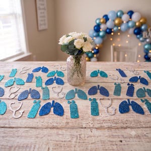 May include: A clear glass vase filled with white roses and eucalyptus leaves sits surrounded by scattered glittery blue paper cutouts shaped like human lungs, oxygen tanks, and stethoscopes, arranged on a rustic light-colored wooden surface.