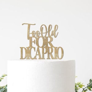May include: A gold glitter cake topper that reads "Too Old For DiCaprio" in a decorative font. The topper is placed on top of a white cake, with greenery in the foreground.
